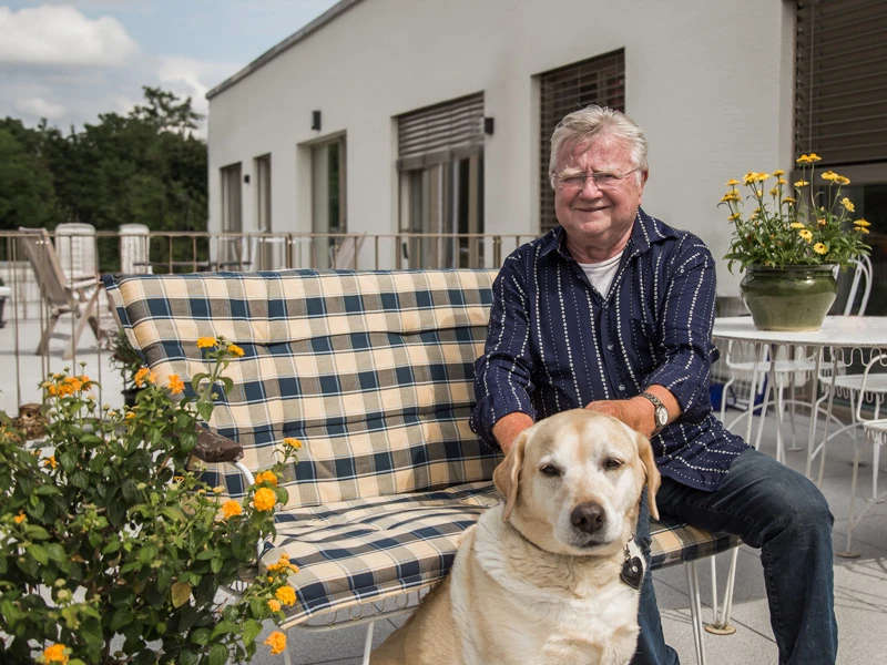 DSW24 – Dresder Servicewohnen GmbH – Mieter Herr Voigt mit Hund Leo Mann sitzt mit seinem Hund auf einer Bank auf einer Terrasse einer Residenz.